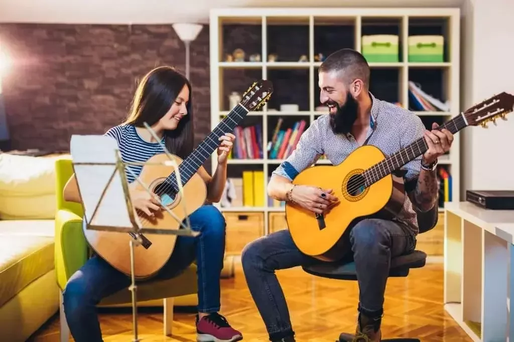 Two people playing guitars and smiling in a cozy living room with bookshelves. A music stand is in the foreground. Warm, joyful scene.