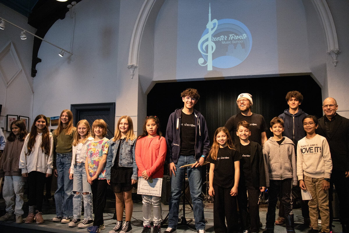 Group of smiling children and adults on stage, diverse outfits, with Greater Toronto Music School logo projected above.