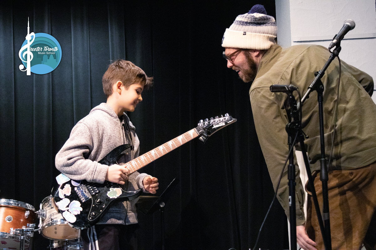 Child plays a sticker-covered electric guitar on stage with a smiling instructor. Logo reads Greater Toronto Music School. Black curtain backdrop.