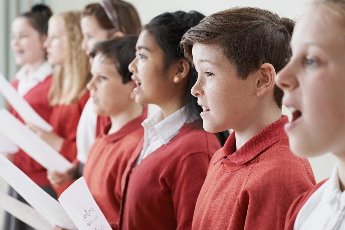 Children in red uniforms sing together, holding sheet music. They appear focused and engaged in a classroom setting.