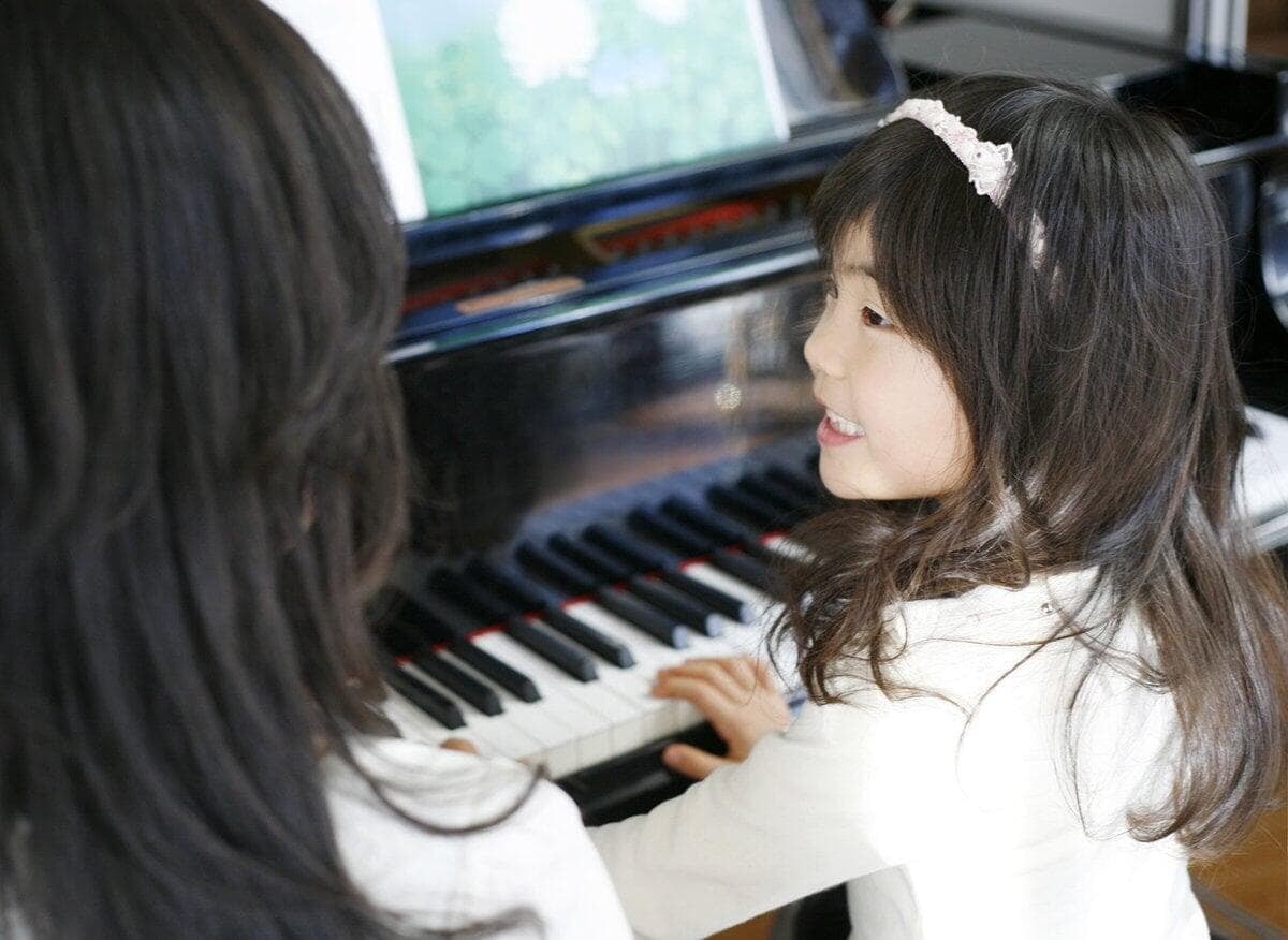 A child plays the piano, smiling as an adult watches closely. The room is bright, and a sheet of music is placed on the piano.