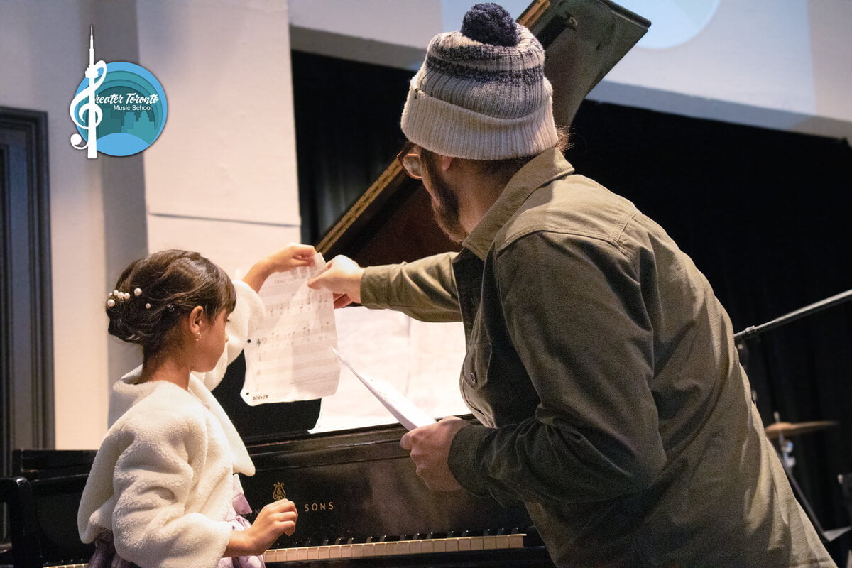 A man helps a young girl with sheet music at a piano. The room has a calm, instructional atmosphere. Logo: Greater Toronto Music School.