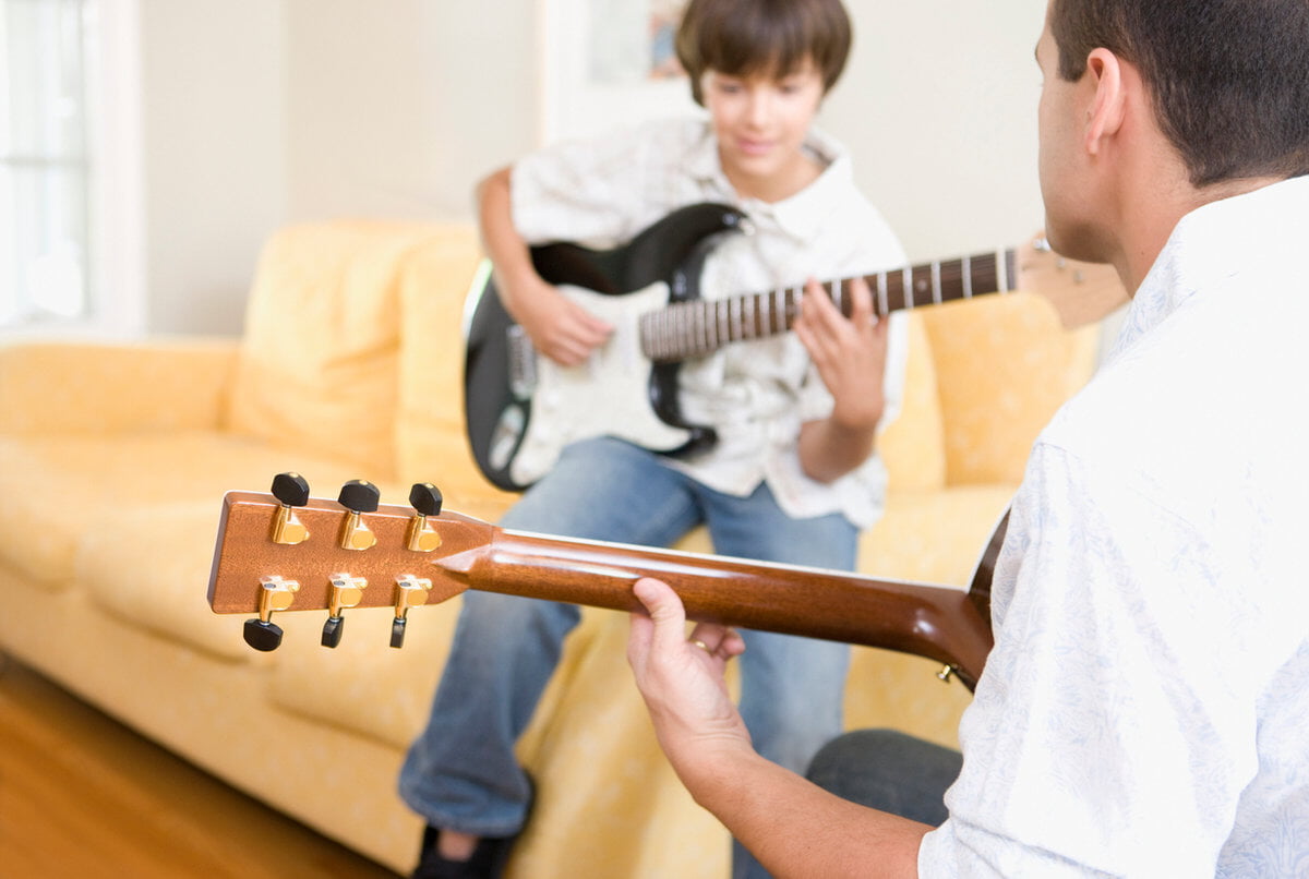 Two people are playing guitars in a cozy room with a yellow couch. One man is guiding a child with a black electric guitar.
