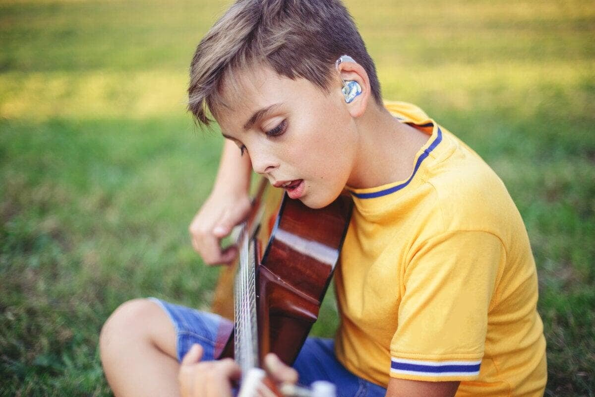 Boy with hearing aid plays guitar in a grassy park, wearing a yellow shirt. Focused expression, serene setting.