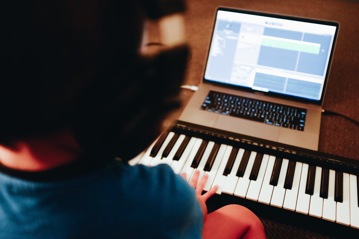 Person with headphones plays a keyboard, focused on a laptop displaying music software. Warm lighting creates a creative mood.