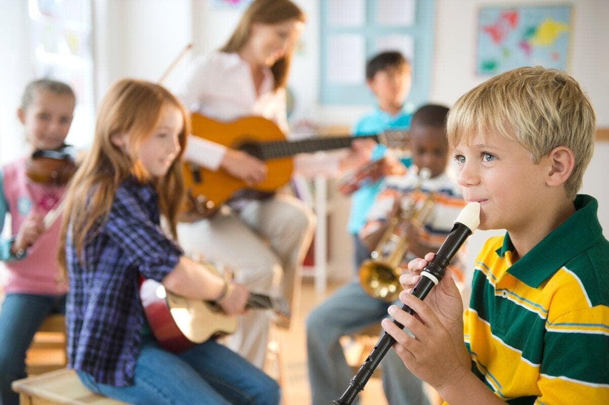 Children in a classroom play various instruments, including a recorder and violin, led by a teacher with a guitar. Bright and cheerful setting.