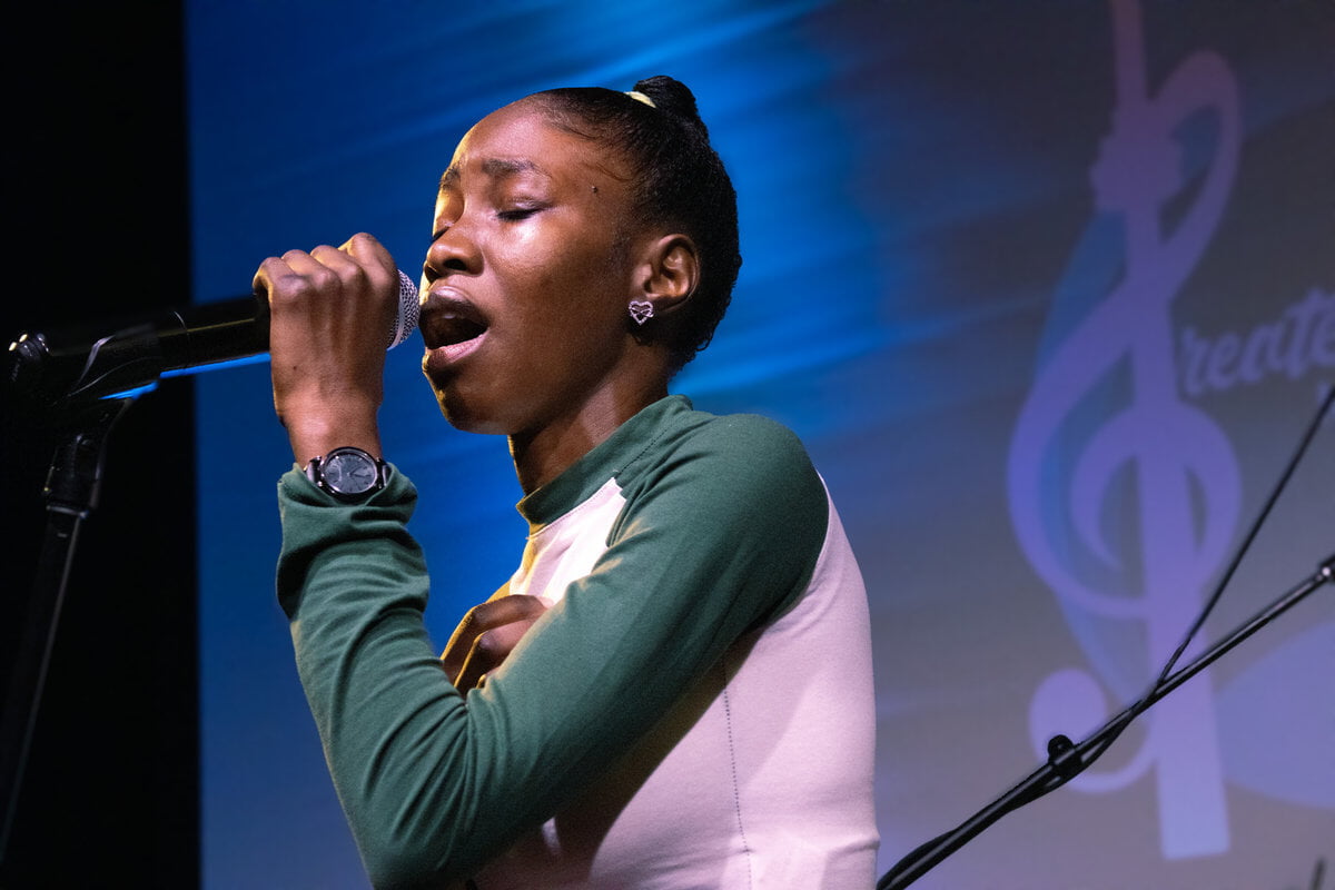 Singer with closed eyes, wearing green and white, emotionally performs on stage. Blue-lit background with musical note design.