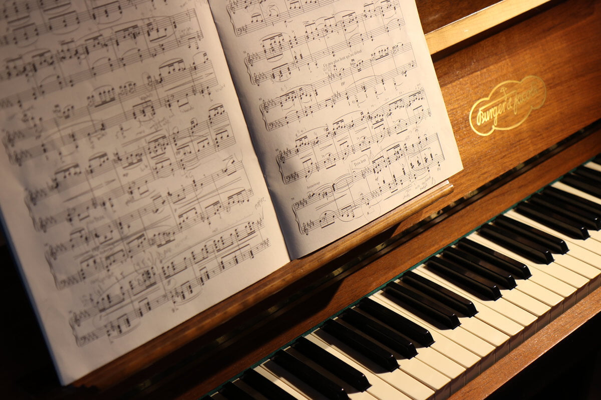 Sheet music on a wooden piano with visible keys and soft lighting. The piano brand logo is visible, creating a warm, musical ambiance.