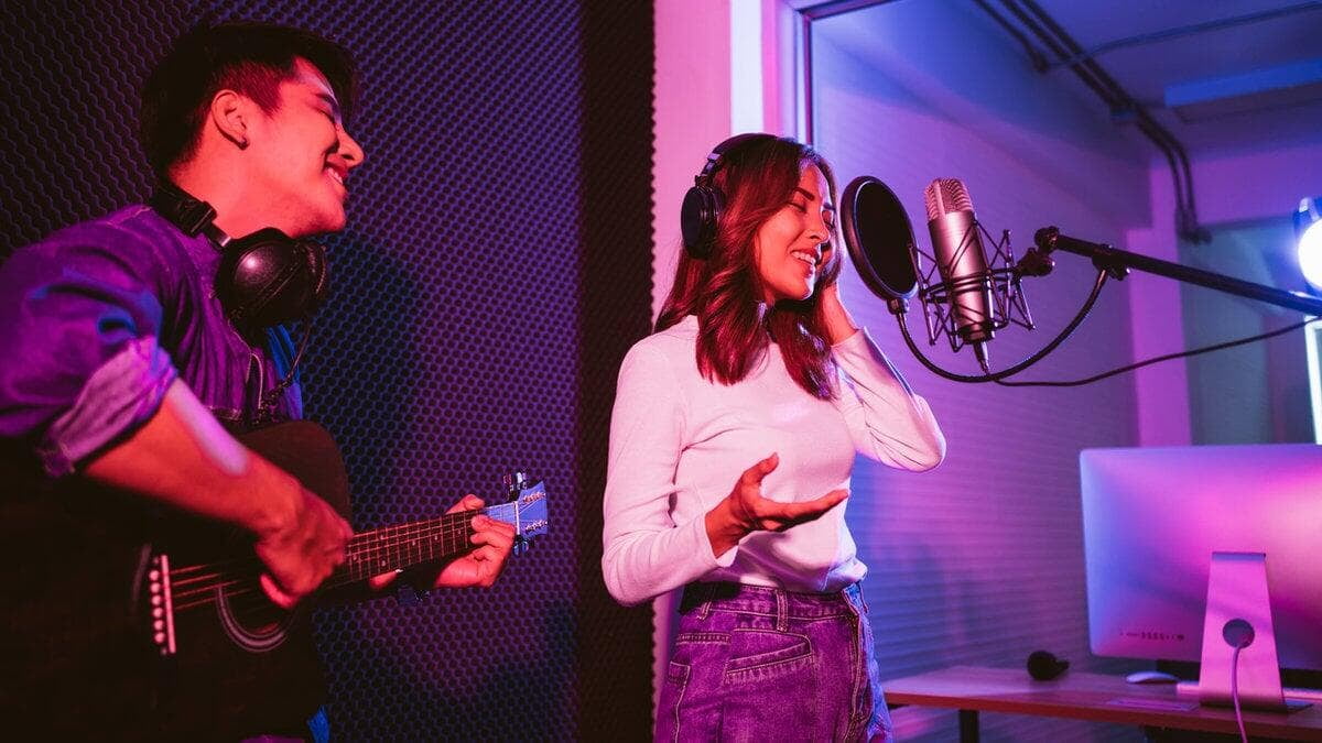 Singers in a studio: woman sings into a mic, man plays guitar. Purple lighting, acoustic paneling, and monitor screen in background.